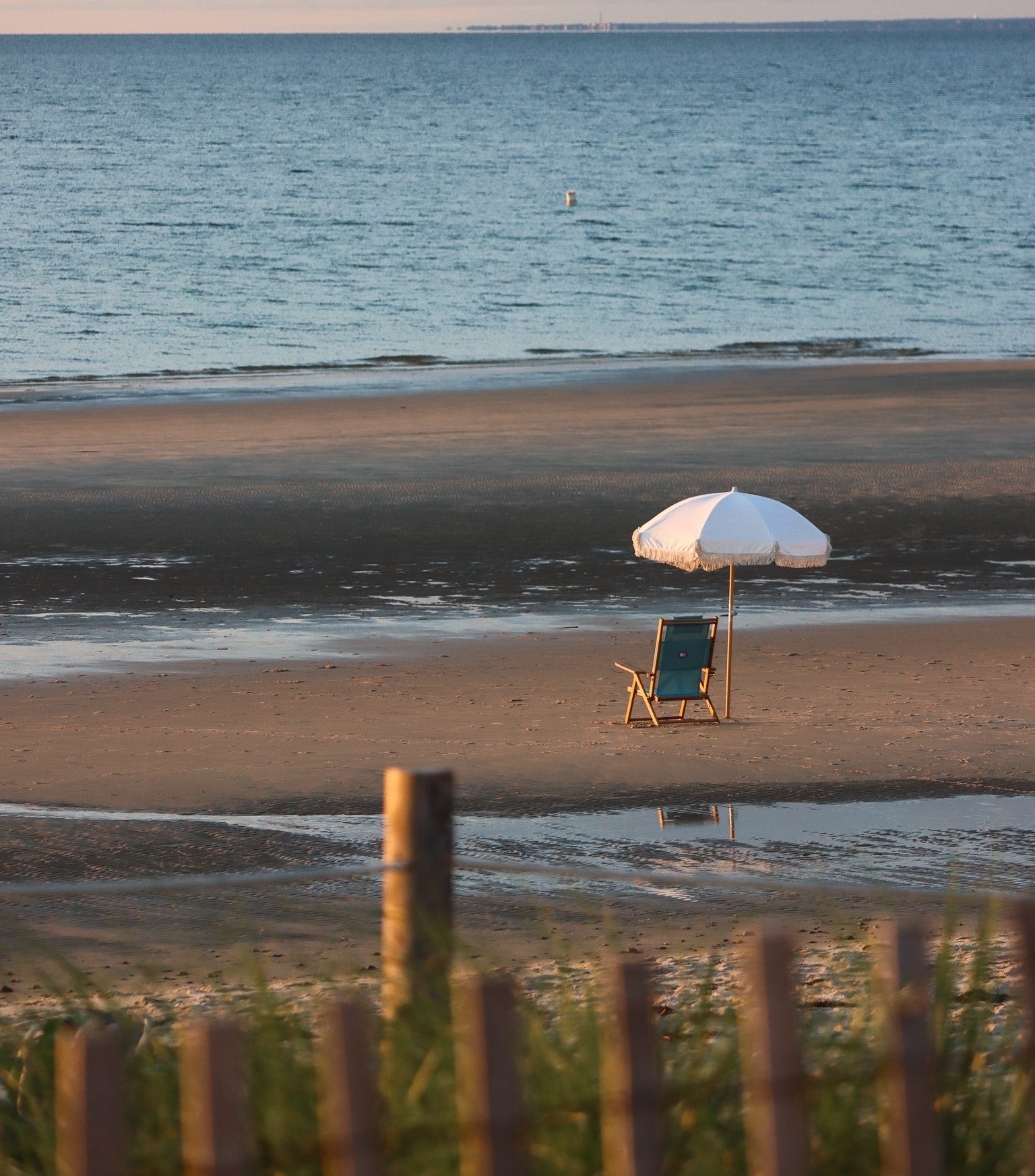 One of our luxury teak beach chair sits under a premium beach umbrella on a sandy beach, overlooking gentle waves.